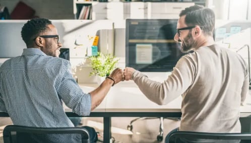 Two men joining fists at a desk in an office, symbolizing a successful business agreement or partnership