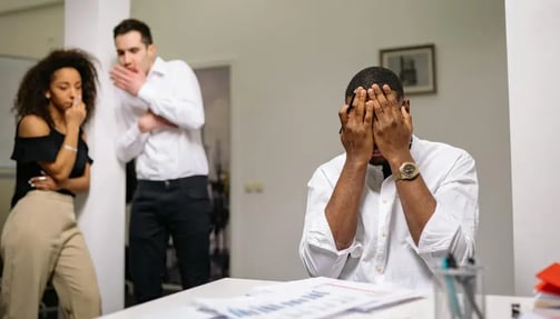 A man stands in front of a desk, covering his face with his hands, with two people at the back covering their mouth and whispering