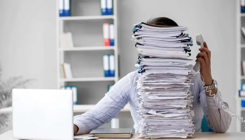 Office worker buried under a towering stack of paperwork while multitasking on the phone.