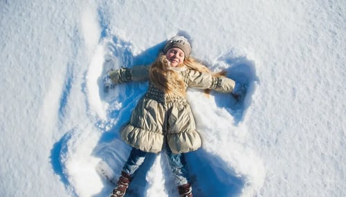 A girl in a coat and hat joyfully creates a snow angel in a winter landscape, surrounded by fresh, white snow