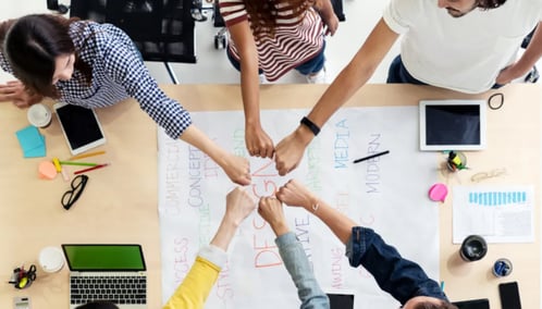 A diverse group of individuals join hands around a table, symbolizing unity, teamwork, and collaboration.